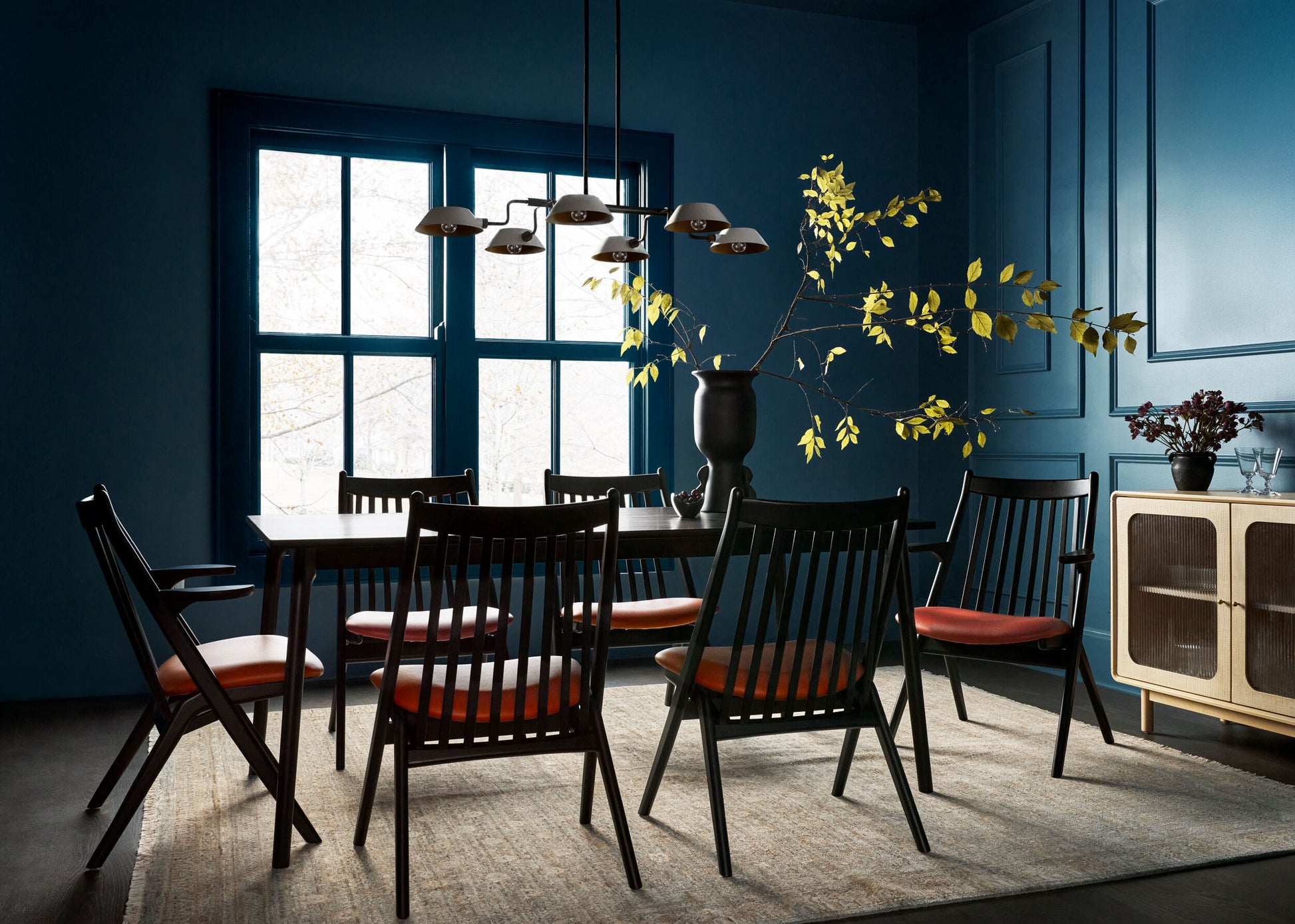 Modern dining room with a black wood dining table, spindle-back chairs, blue walls, and warm lighting.