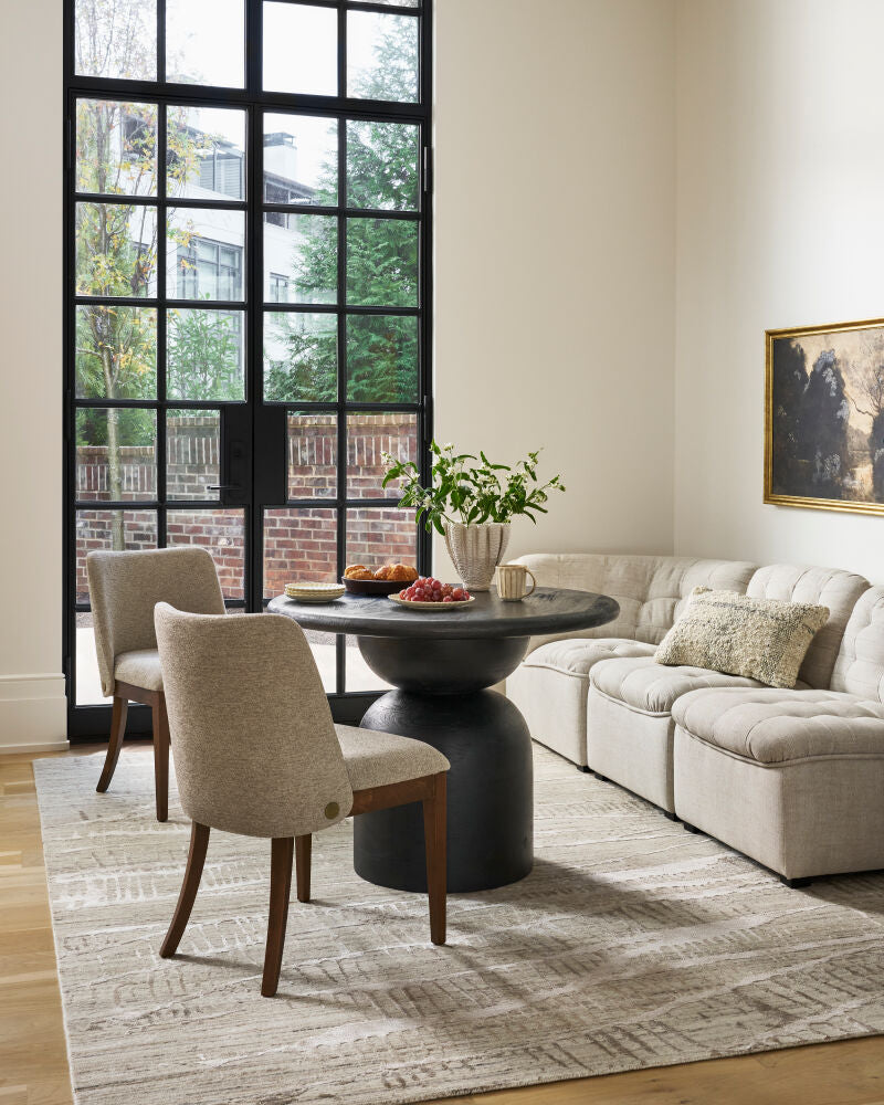 Black mango wood pedestal dining table in a bright space with beige chairs, a tufted sectional, and decor.
