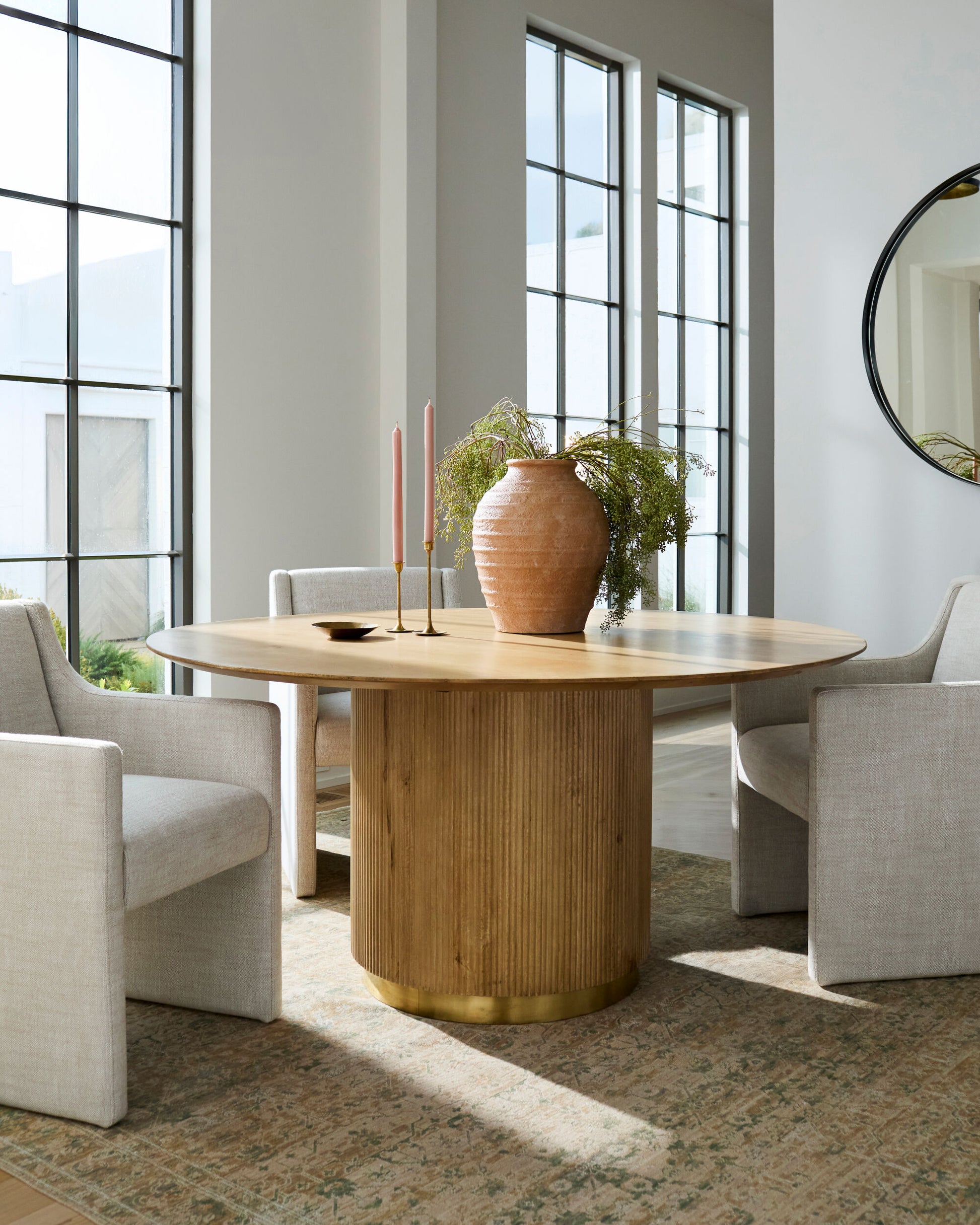 Round mango wood dining table with fluted pedestal, brass accents, and beige chairs in bright room.