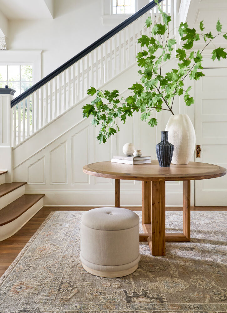 Round Acacia wood dining table with a warm finish, beige ottoman, and decor on a vintage rug.