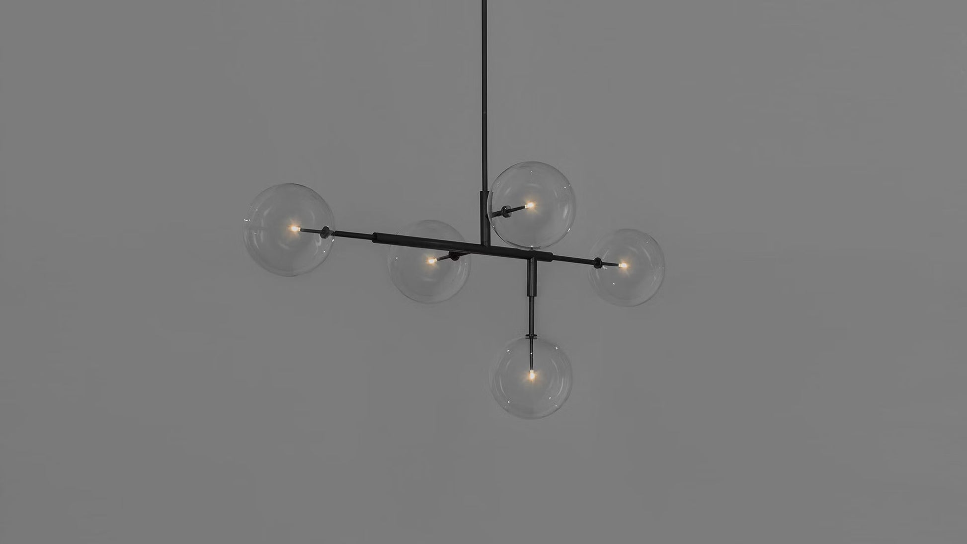 Black gunmetal chandelier with five glass globes, illuminated against a gray wall.