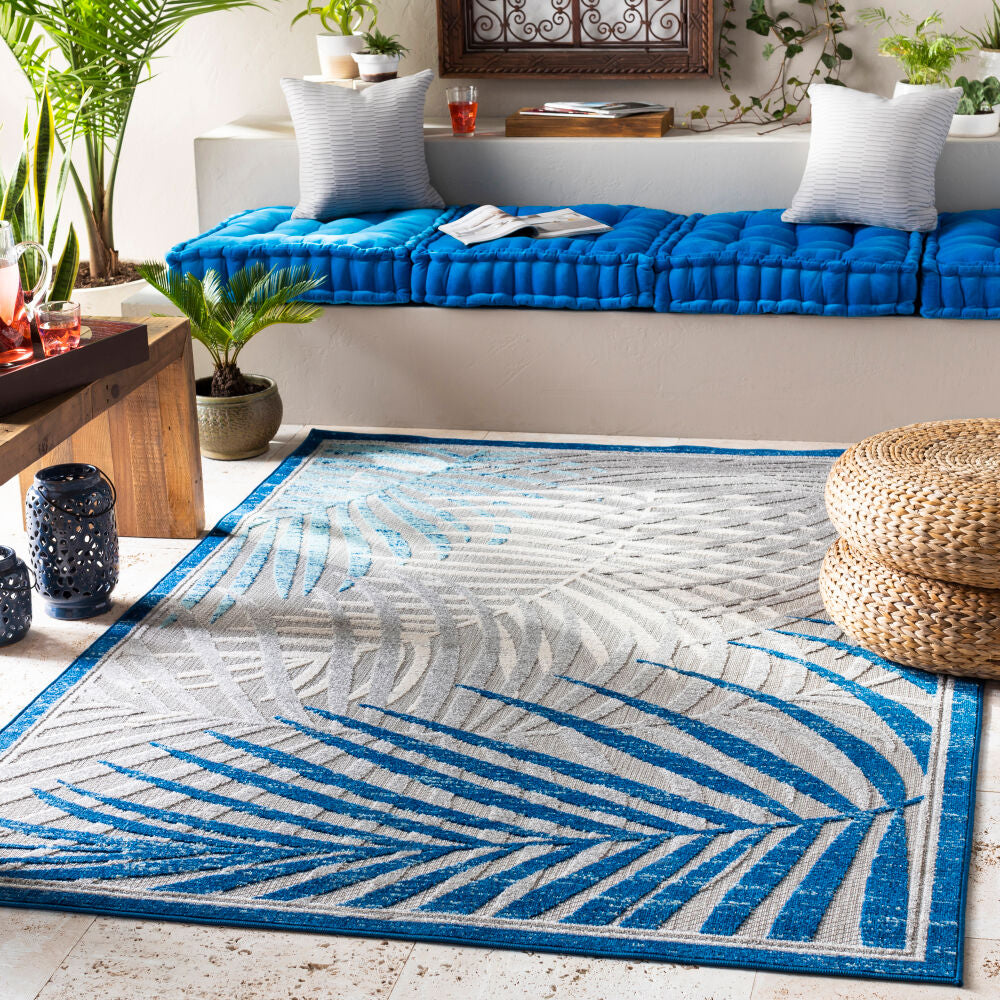 Blue and grey tropical palm leaf rug displayed in an indoor seating area with a vibrant blue cushion and wicker ottomans.