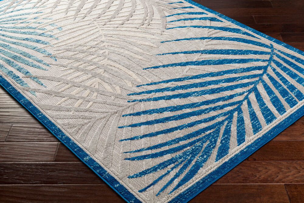 Close-up of tropical area rug with blue palm leaf patterns against a gray textured background on wooden flooring.