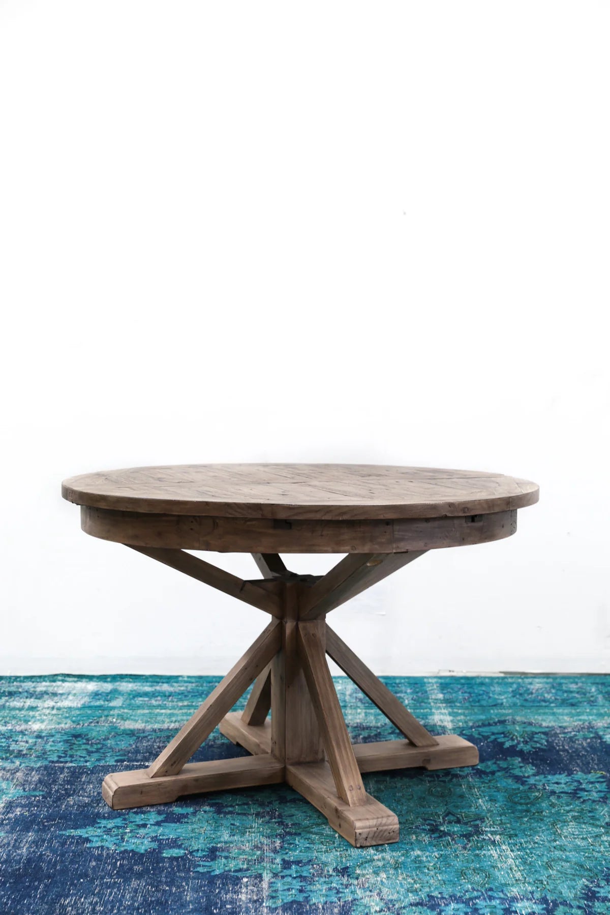 Irish Coast round extension dining table with a rustic light brown finish, displayed on a blue-patterned rug.