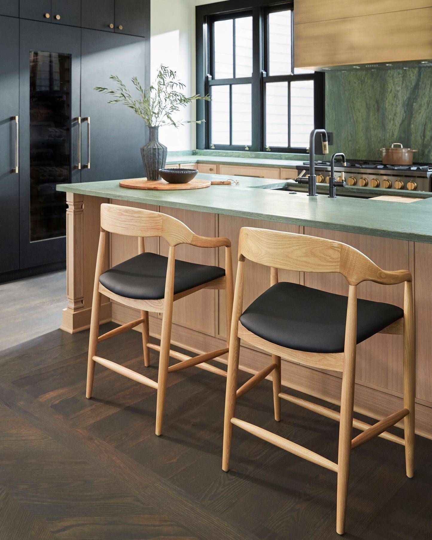 Natural ash wood bar stools with black faux leather seats at a modern kitchen island with green stone countertops.